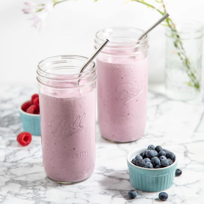 Two light purple smoothies with a bowl of blueberries.