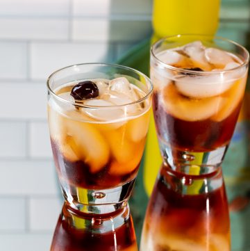 Two dark brown cocktails garnished with a cherry sitting on a mirrored surface.