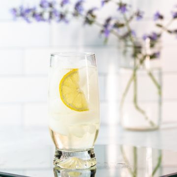 Light yellow cocktail with a lemon wedge in front of a vase filled with purple flowers.