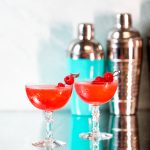 Two Bright red cocktails garnished with raspberries in front of cocktail shakers.