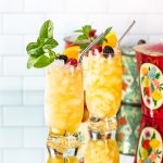 Two bright yellow cocktails garnished with fruit, mint and a metal straw.