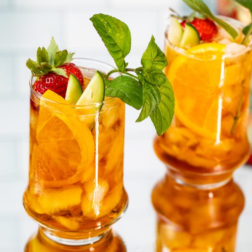 Two cocktails garnished with an orange wheel, strawberry, cucumber and mint.