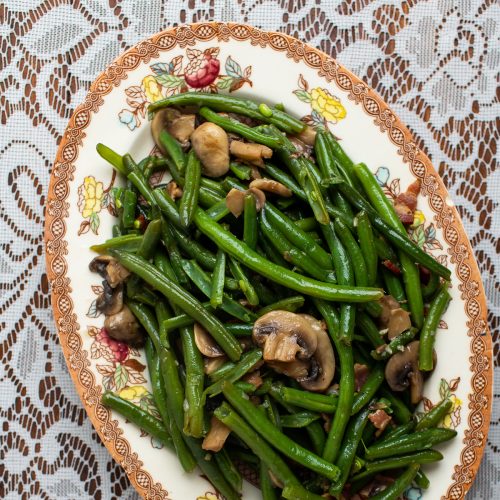 Platter of green beans with mushrooms and bacon on a lace tablecloth.